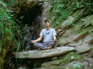 Man Sitting Cross Legged on Rock by Waterfall and Meditating